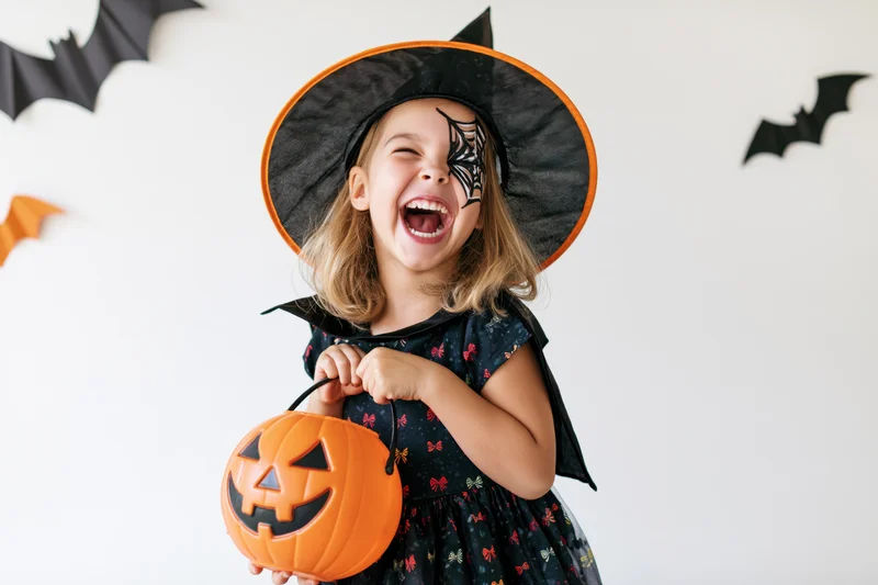 Young girl in witch costume laughing while holding a pumpkin bucket indoors