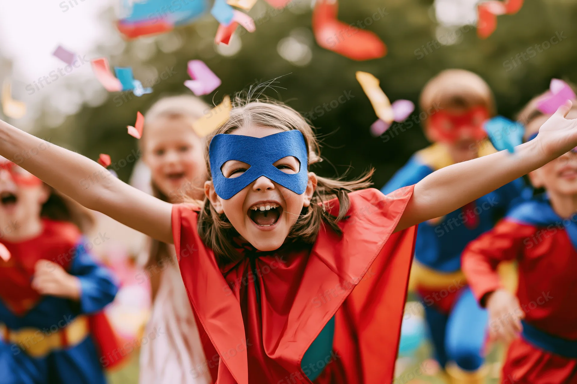 Young girl in superhero costume with red cape and blue mask laughing with arms outstretched outdoors among friends during a festive celebration