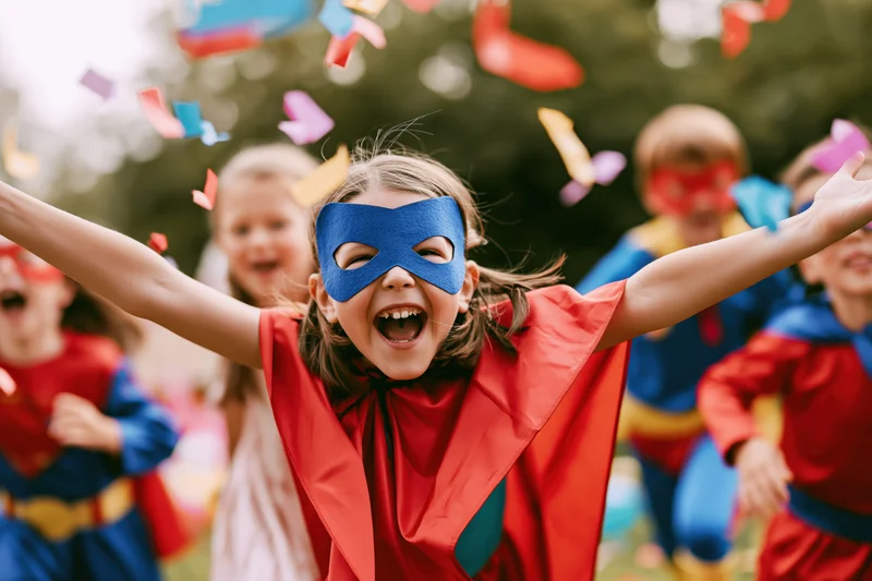 Young girl in superhero costume with red cape and blue mask laughing with arms outstretched outdoors among friends during a festive celebration