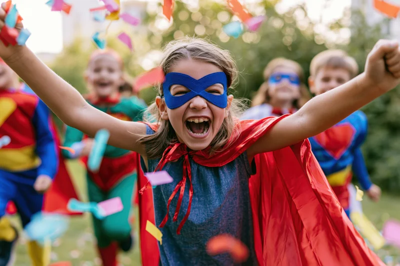 Girl in superhero costume with red cape and blue mask laughing with arms raised outdoors among children in costumes