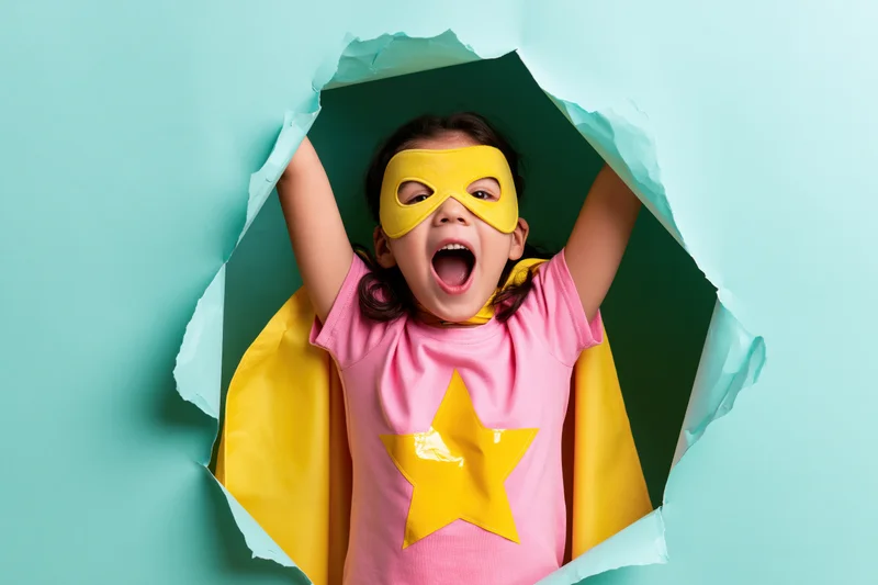 Girl in superhero costume with mask and cape emerging through torn paper hole
