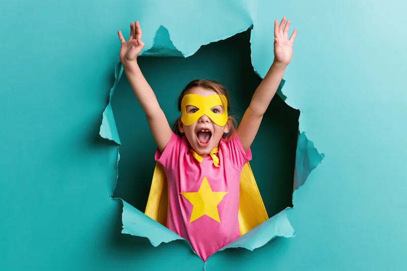 Girl in superhero costume with arms raised emerging from torn paper hole