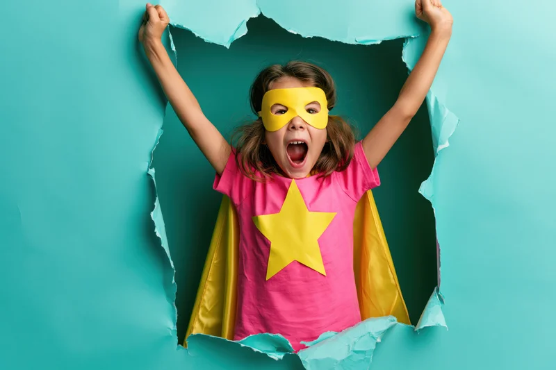 Girl in superhero costume tearing through paper wall with arms raised and mouth open