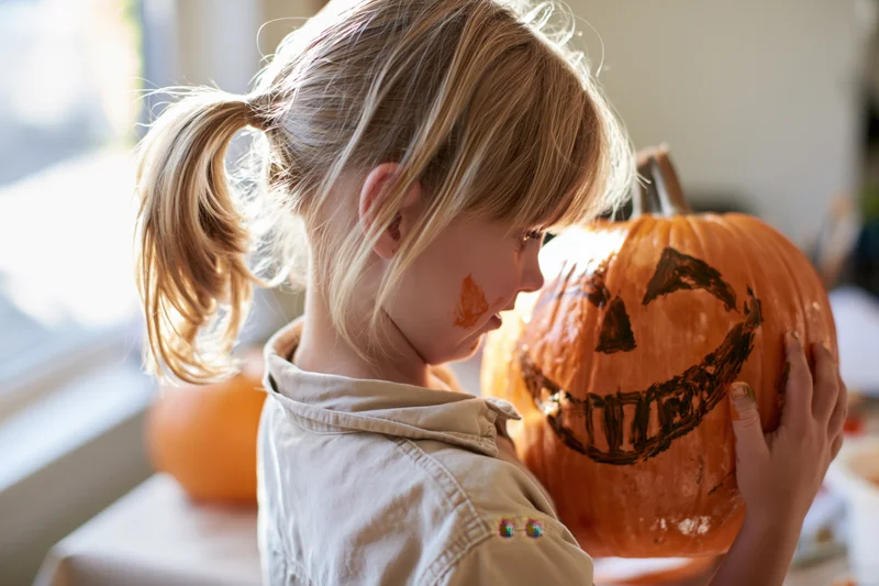Young girl painting a jack-o-lantern face with marker indoors near a window