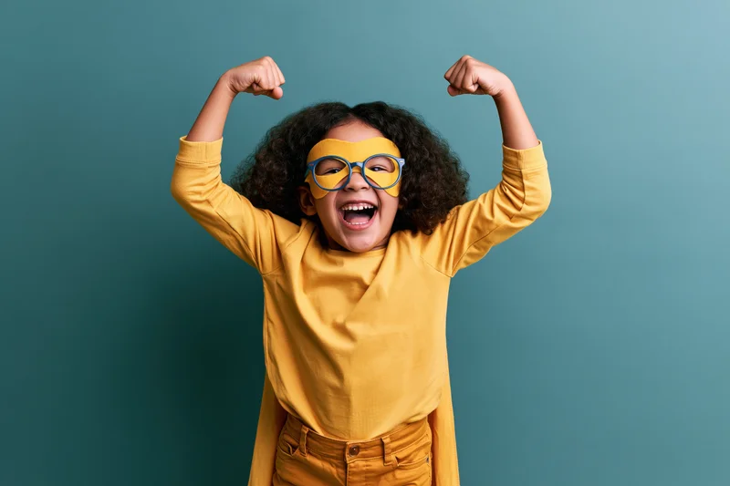 Young girl in superhero costume flexing arms with joyful expression against teal background