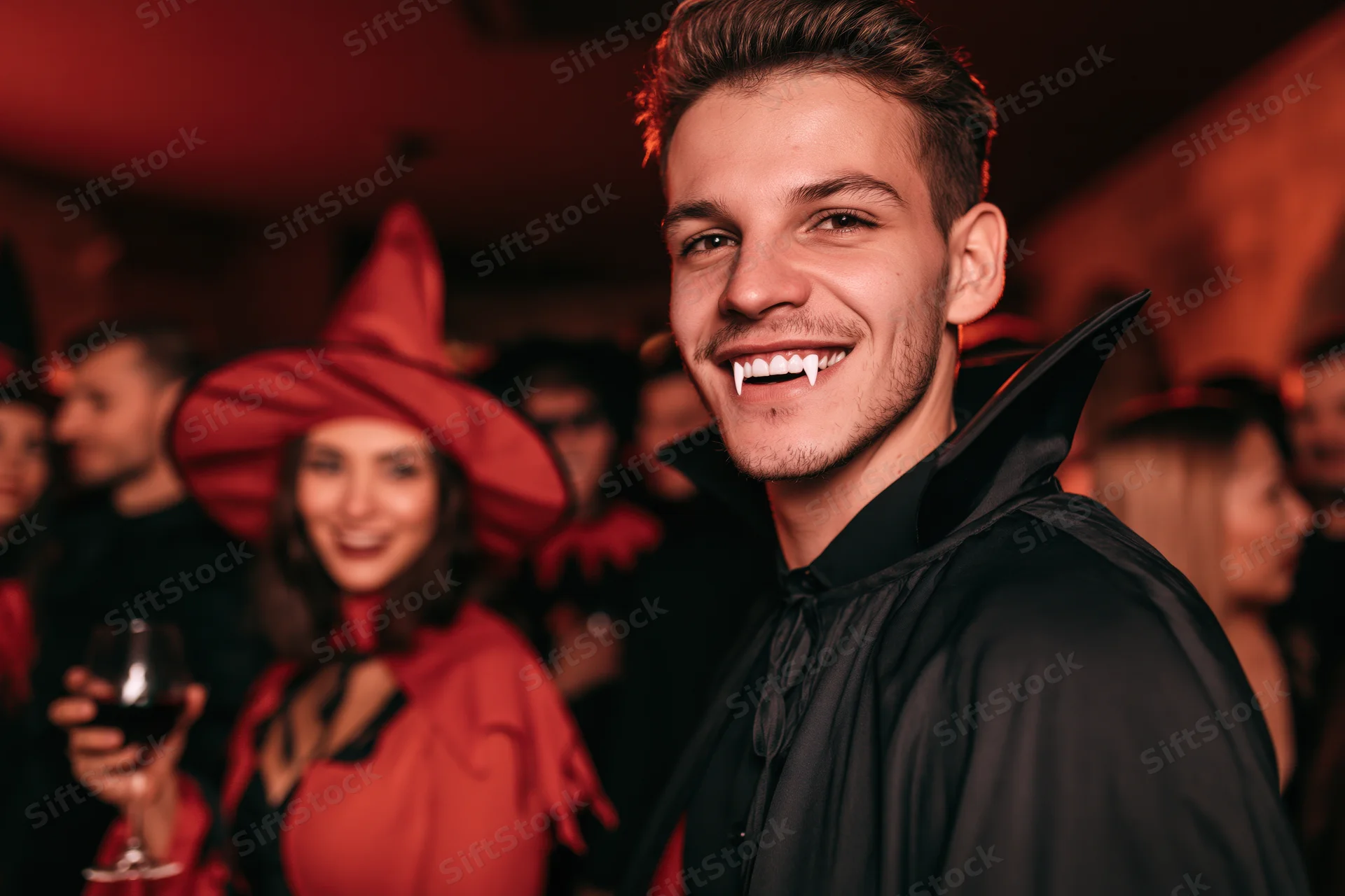 Man in vampire costume smiling at camera at halloween party with woman in witch hat holding wine glass in background
