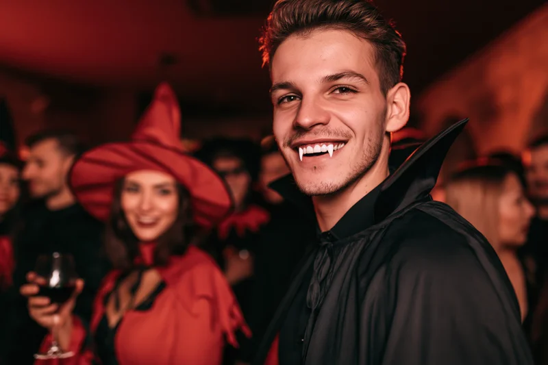 Man in vampire costume smiling at camera at halloween party with woman in witch hat holding wine glass in background