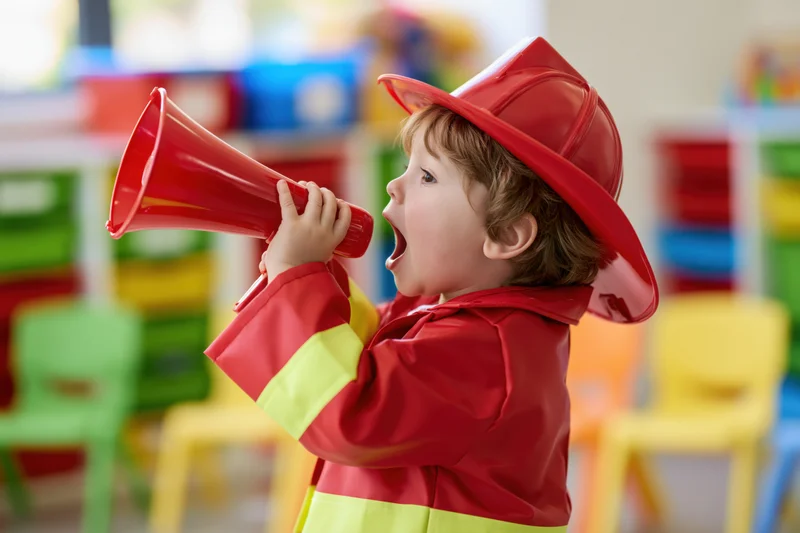 Toddler boy in red firefighter costume shouting into megaphone in playroom