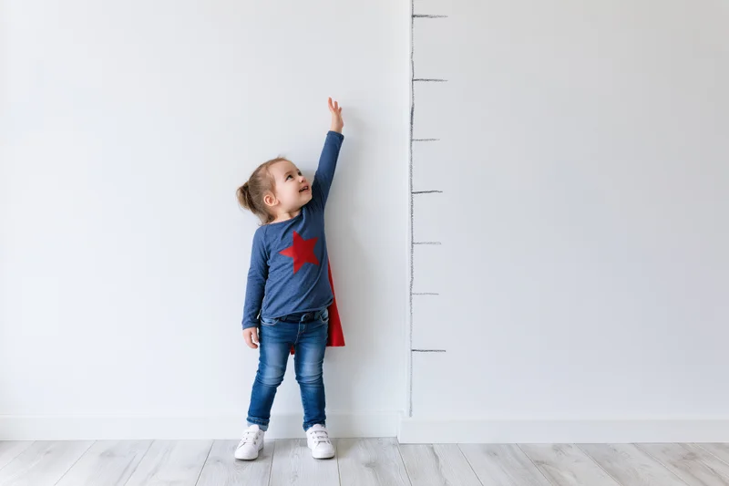 Young girl standing against white wall reaching up to mark her height on a growth chart