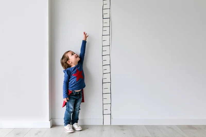 Toddler girl standing against white wall reaching up to height measurement chart while wearing superhero costume