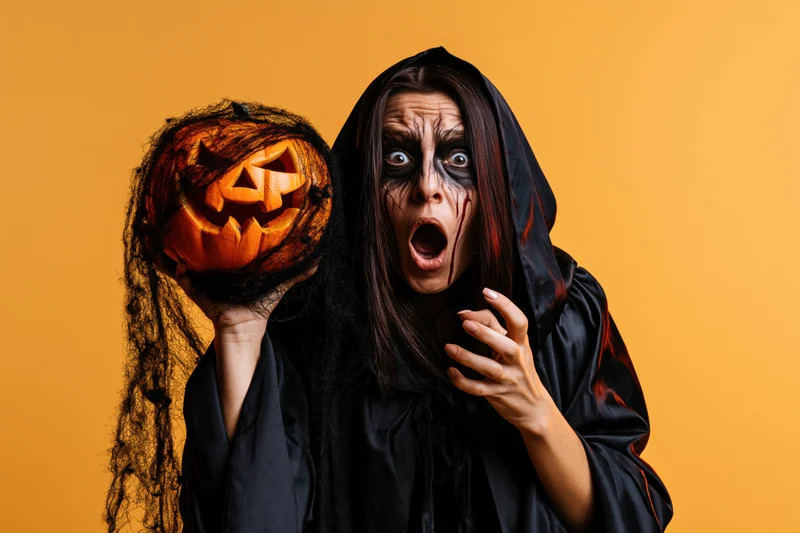 Woman in black witch costume holding carved pumpkin with surprised expression against orange background