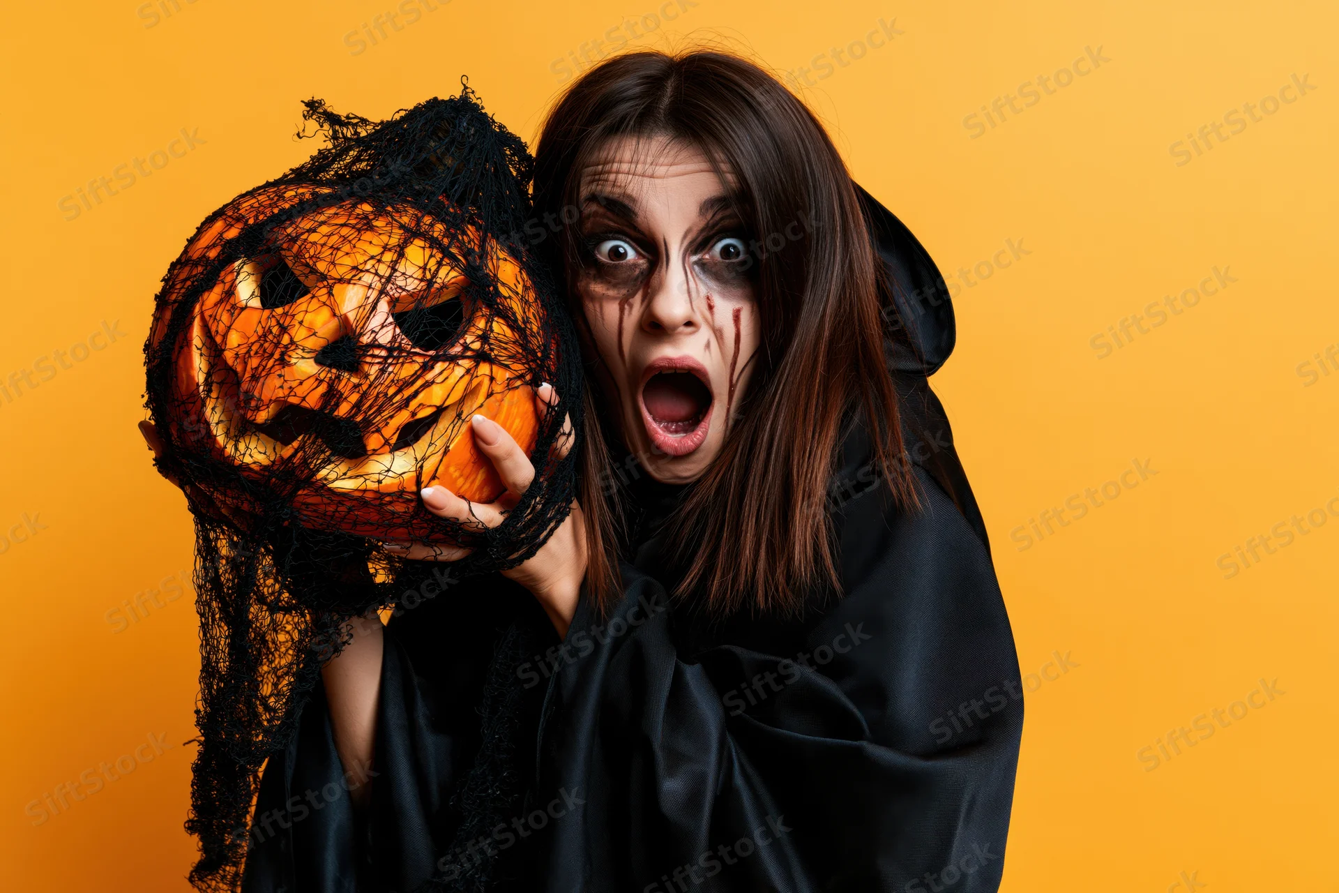 Woman in black witch costume holding carved pumpkin with netting, orange background