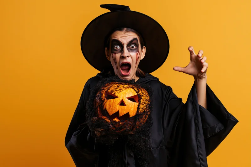 Woman in witch costume holding jack-o-lantern with dramatic expression against orange background