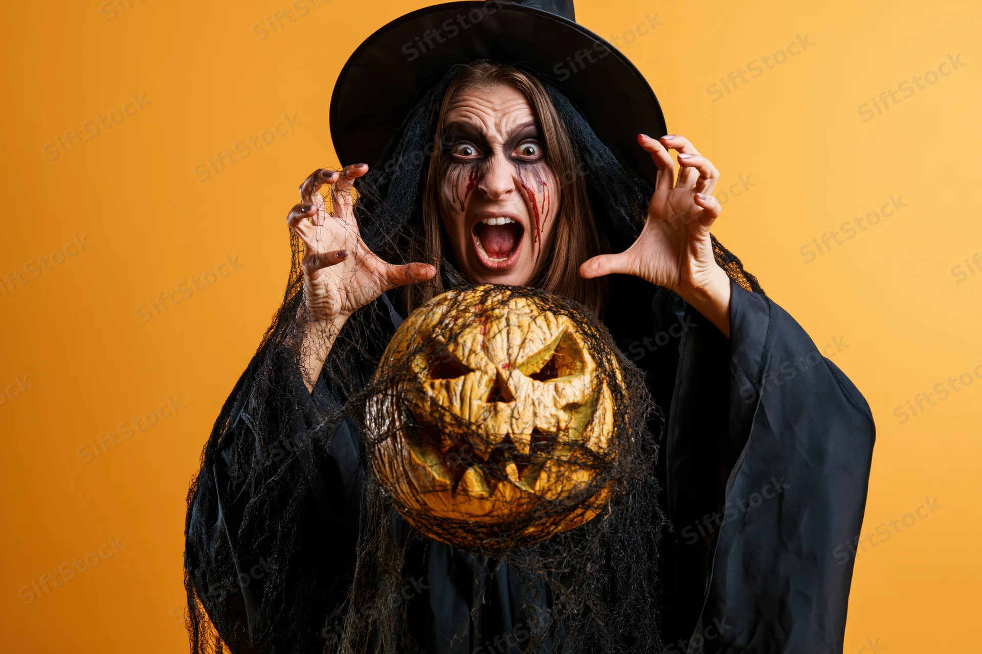Woman in witch costume screaming with carved pumpkin in front of orange background