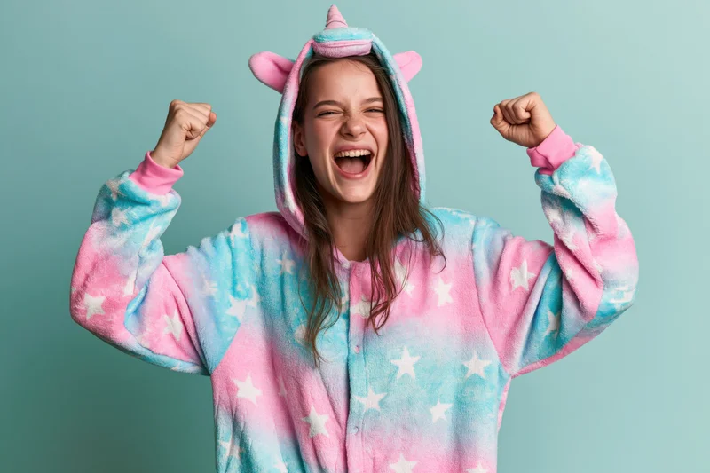 Young woman in a unicorn kigurumi onesie celebrating with raised fists against a pastel background