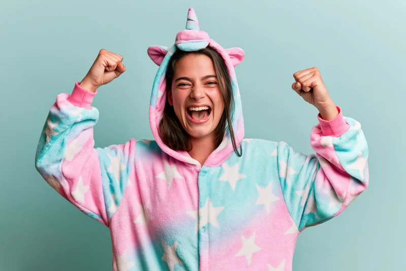 Woman in unicorn kigurumi onesie cheering with raised fists against light blue background