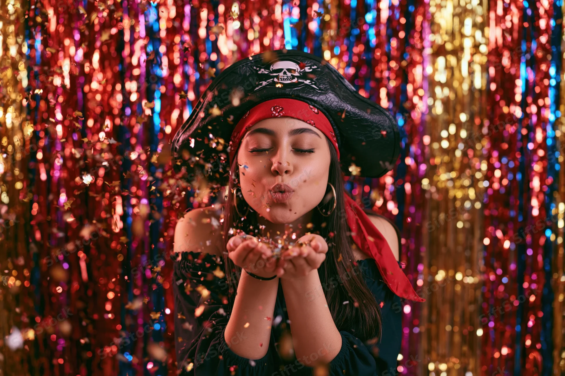 Woman in pirate costume blowing confetti from her cup in front of colorful tinsel backdrop