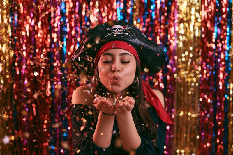 Woman in pirate costume blowing confetti from her cup in front of colorful tinsel backdrop