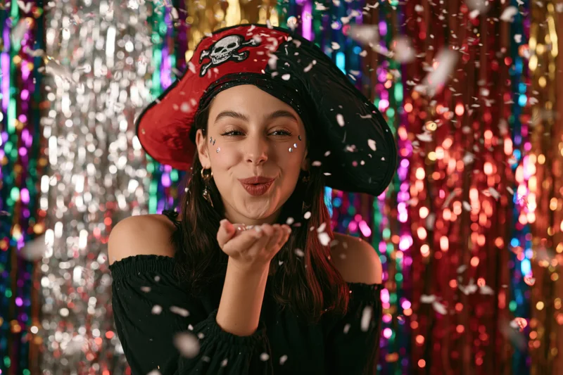 Young woman in pirate costume blowing confetti in front of colorful tinsel backdrop