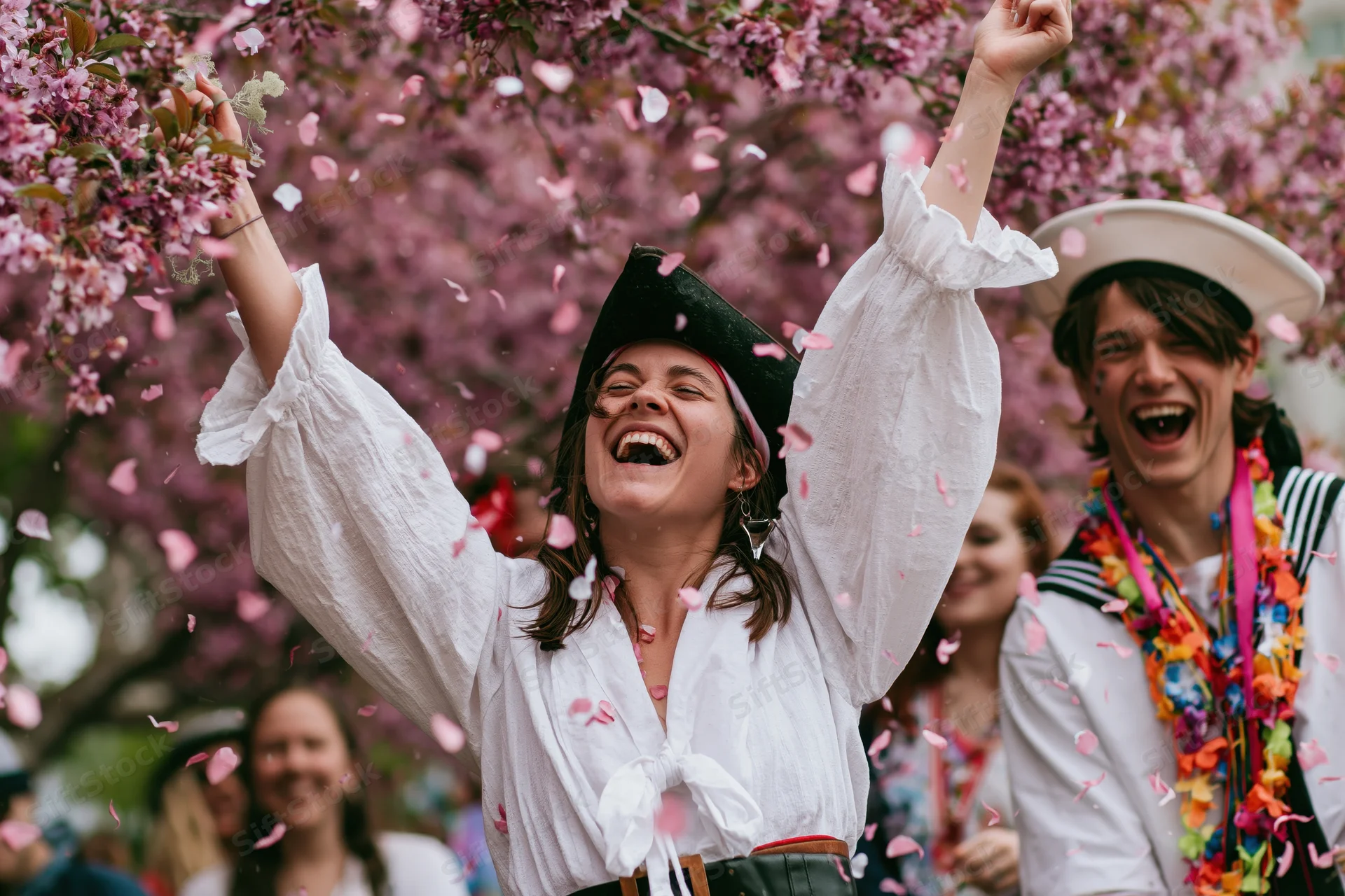 Young woman in pirate costume laughing with arms raised under cherry blossoms at outdoor festival