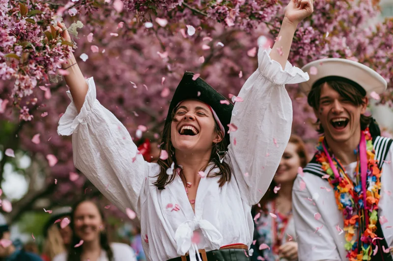 Young woman in pirate costume laughing with arms raised under cherry blossoms at outdoor festival