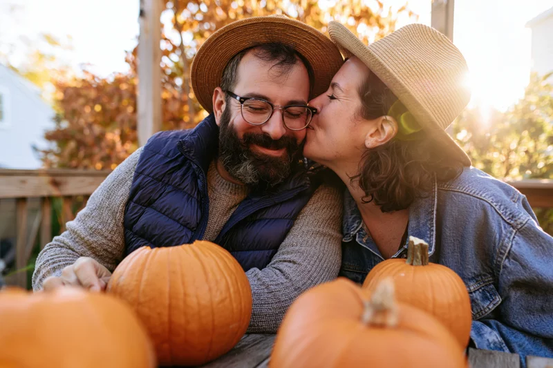 Woman kissing man on the cheek beside pumpkins on a porch