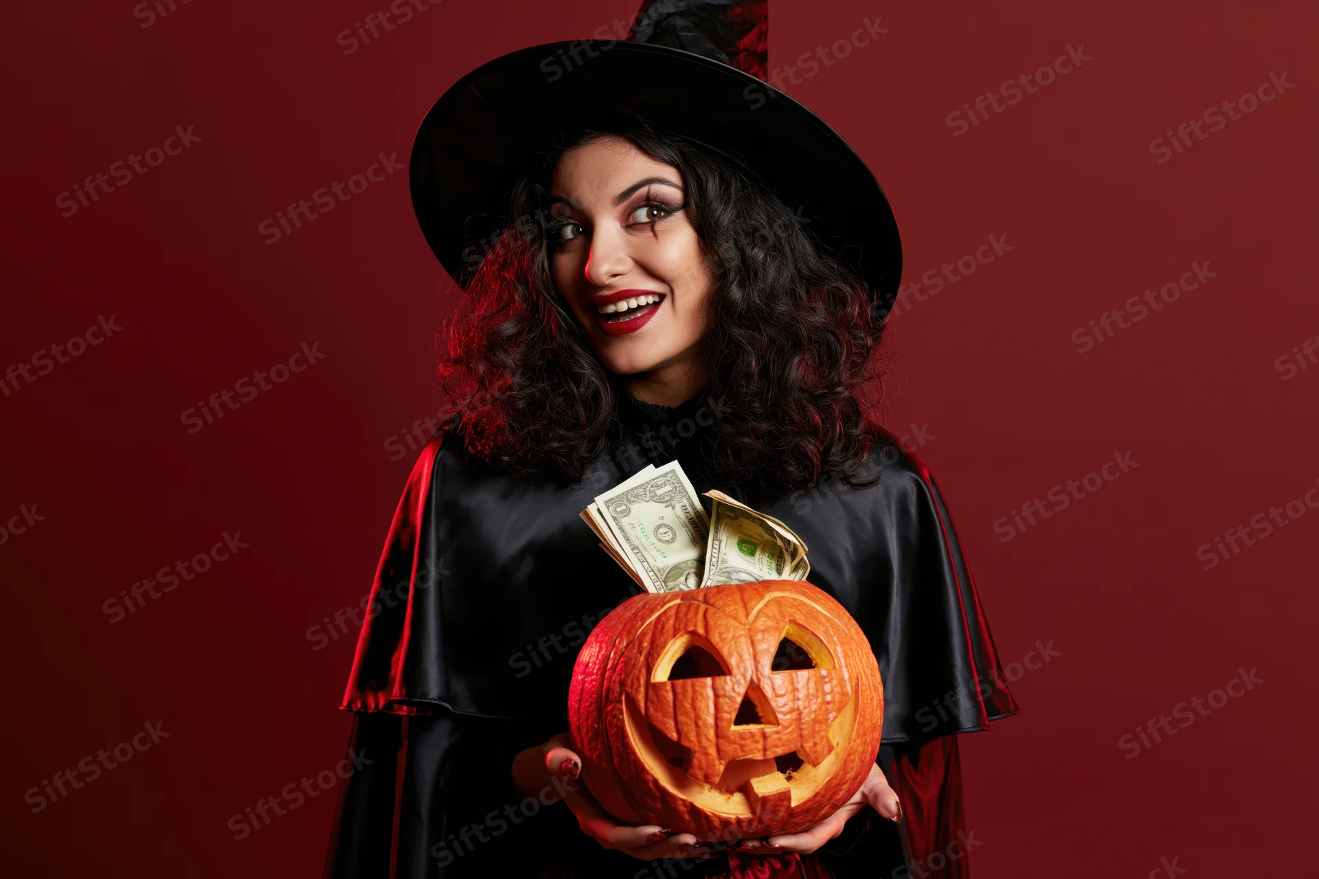 Woman in witch costume holding carved pumpkin with money inside, studio portrait against red background