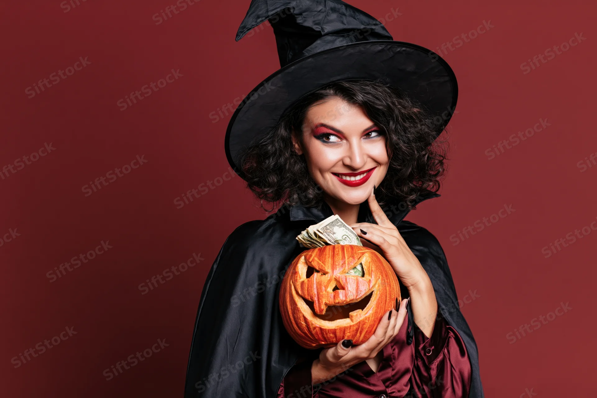 Woman in witch costume holding carved pumpkin with money inside, smiling against red background