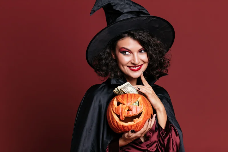 Woman in witch costume holding carved pumpkin with money inside, smiling against red background