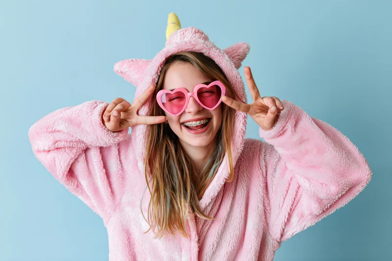 Young woman in pink unicorn onesie making peace signs with heart sunglasses on light blue background