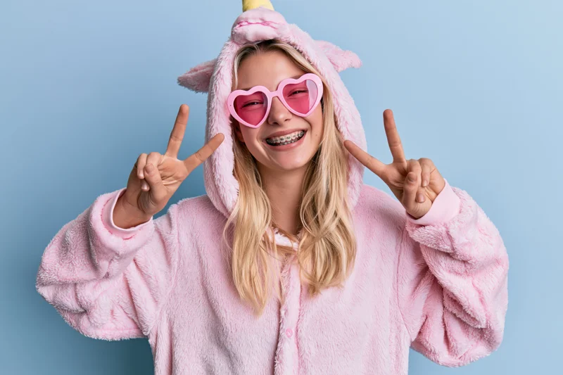 Young woman in pink unicorn onesie making peace signs with heart sunglasses smiling against blue background
