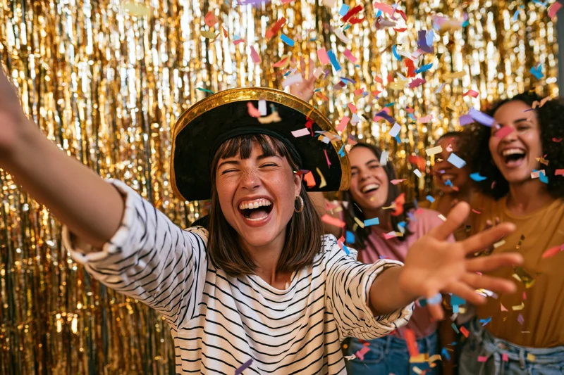 Woman laughing with arms outstretched in front of gold tinsel backdrop as confetti falls around her and friends