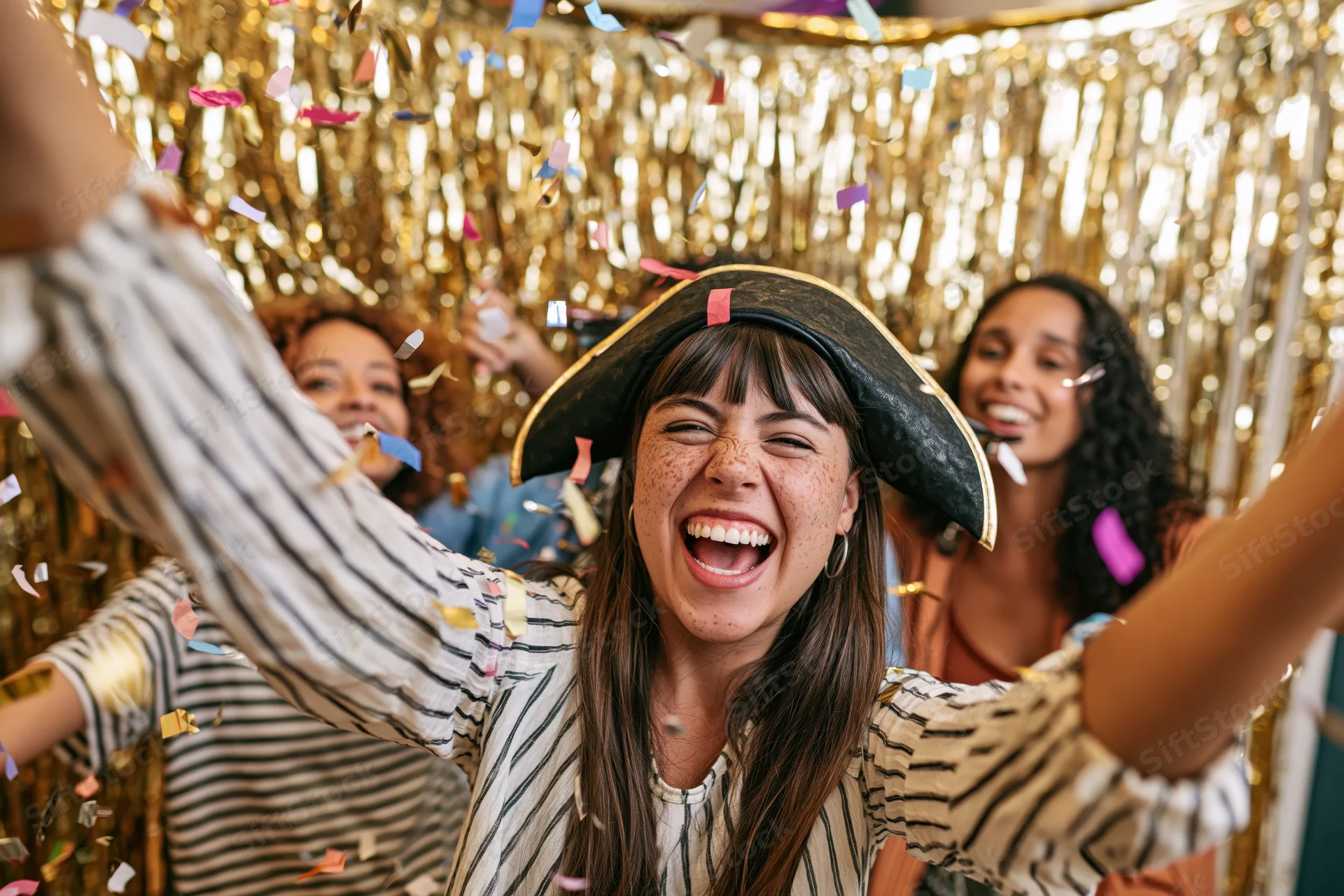 Young woman laughing with arms raised in front of gold tinsel backdrop during a festive party with friends and falling confetti