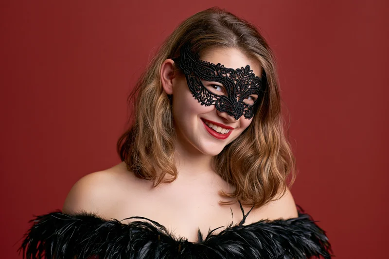 Young woman smiling while wearing ornate black lace masquerade mask and feathered off-shoulder dress against red background