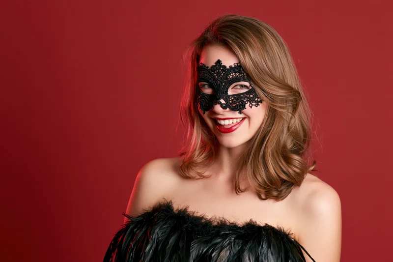 Woman wearing ornate black lace masquerade mask smiling in feathered dress against red background