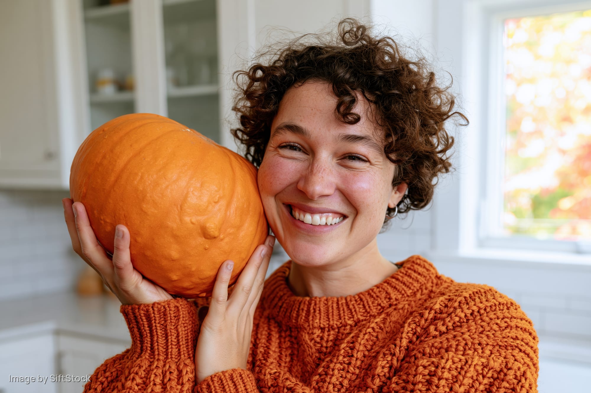 Woman smiling while holding a large orange pumpkin in a kitchen