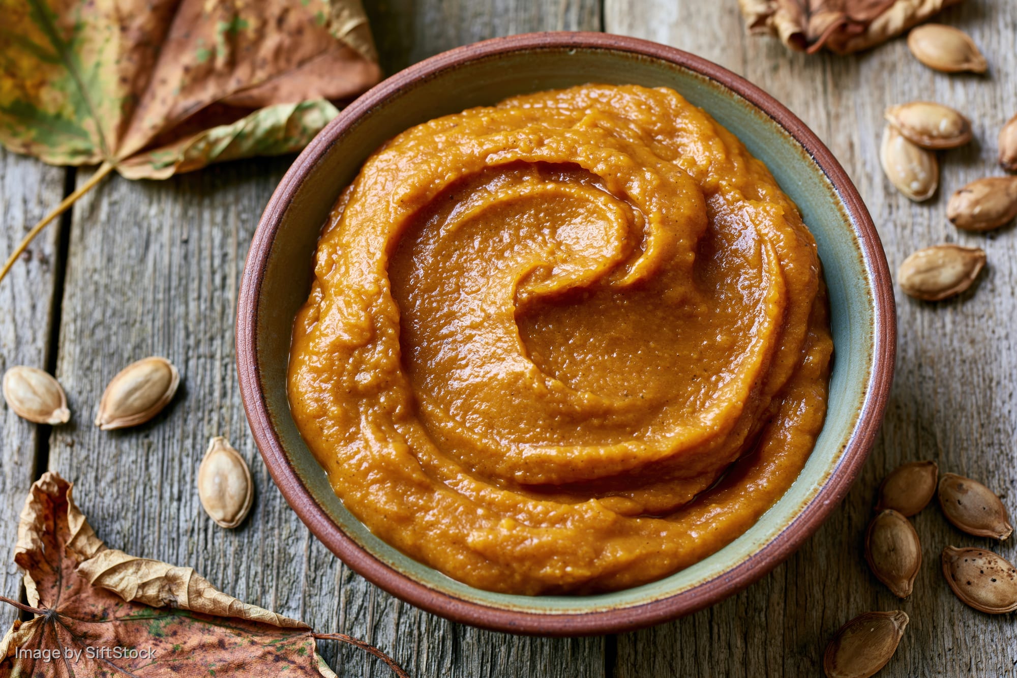 Smooth pumpkin puree in a ceramic bowl on a rustic wooden table surrounded by dried leaves and pumpkin seeds