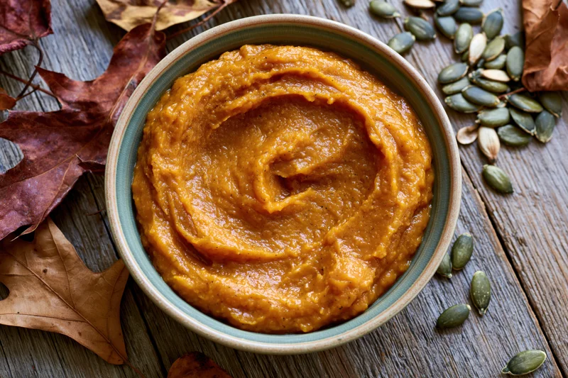 Ceramic bowl of smooth pumpkin puree with swirl pattern on wooden table surrounded by dried leaves and pumpkin seeds