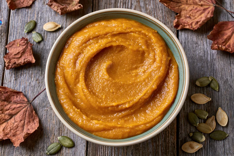 Ceramic bowl filled with smooth pumpkin puree on wooden table surrounded by dried leaves and seeds
