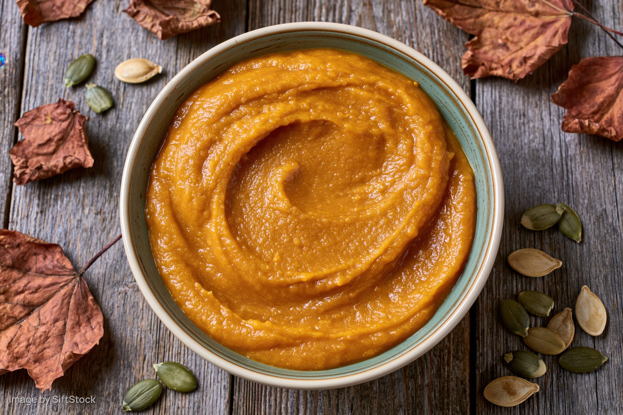 Ceramic bowl filled with smooth pumpkin puree on wooden table surrounded by dried leaves and seeds