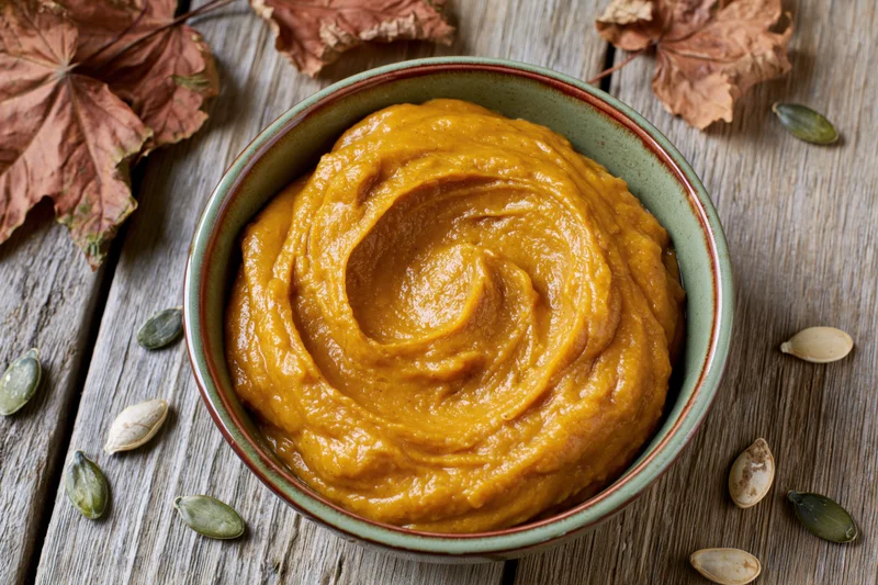 Smooth pumpkin puree in a ceramic bowl on a wooden table surrounded by dried leaves and seeds
