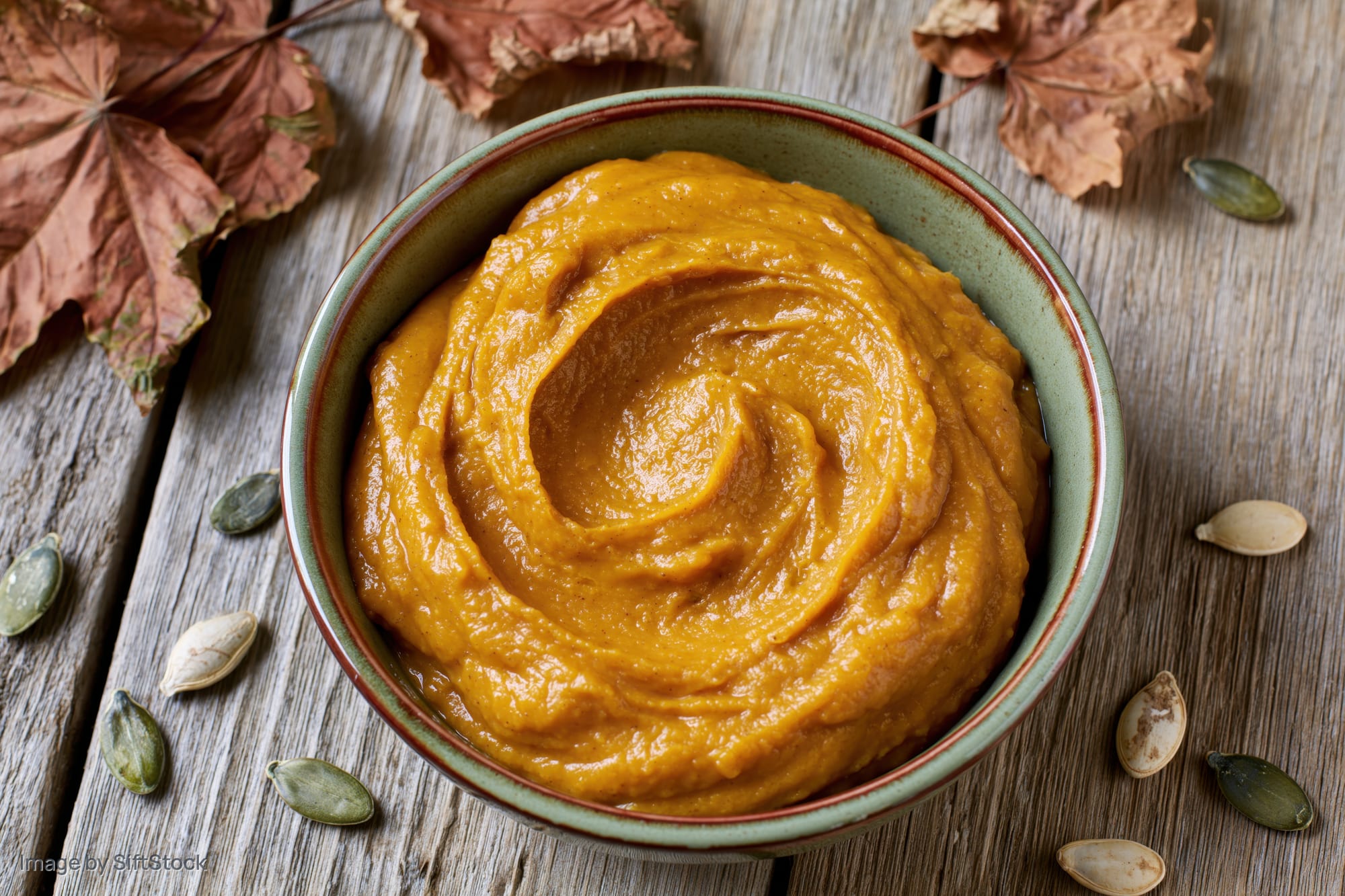 Smooth pumpkin puree in a ceramic bowl on a wooden table surrounded by dried leaves and seeds
