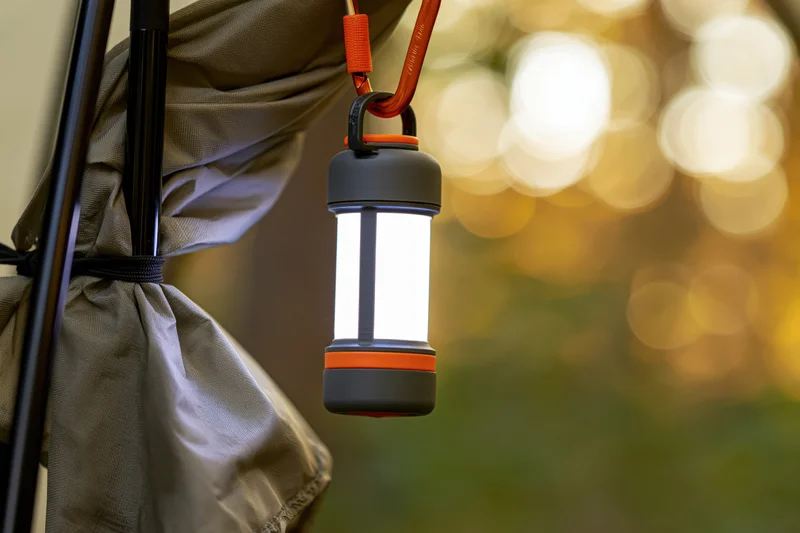 Compact led lantern glowing brightly while hanging from a backpack strap near a tent at dusk