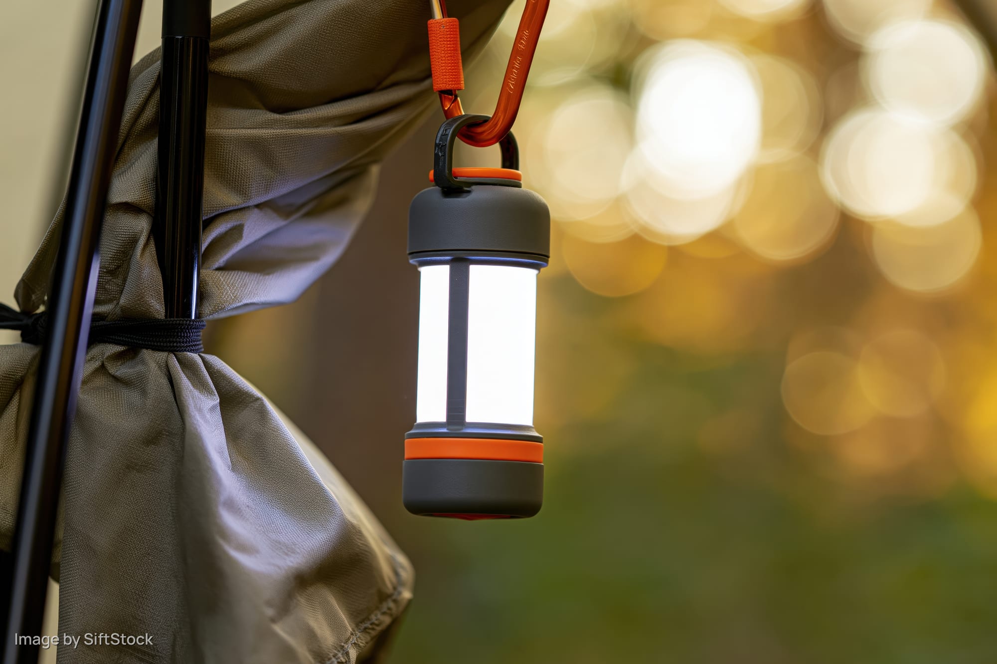 Compact led lantern glowing brightly while hanging from a backpack strap near a tent at dusk