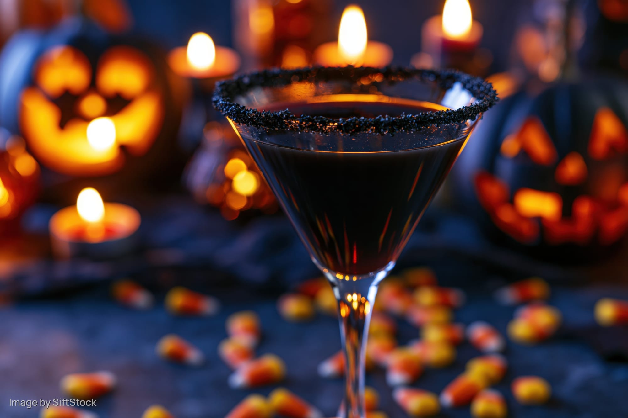 Dark cocktail in a martini glass with sugared black rim surrounded by glowing jack-o-lanterns and candy corn on a dark table