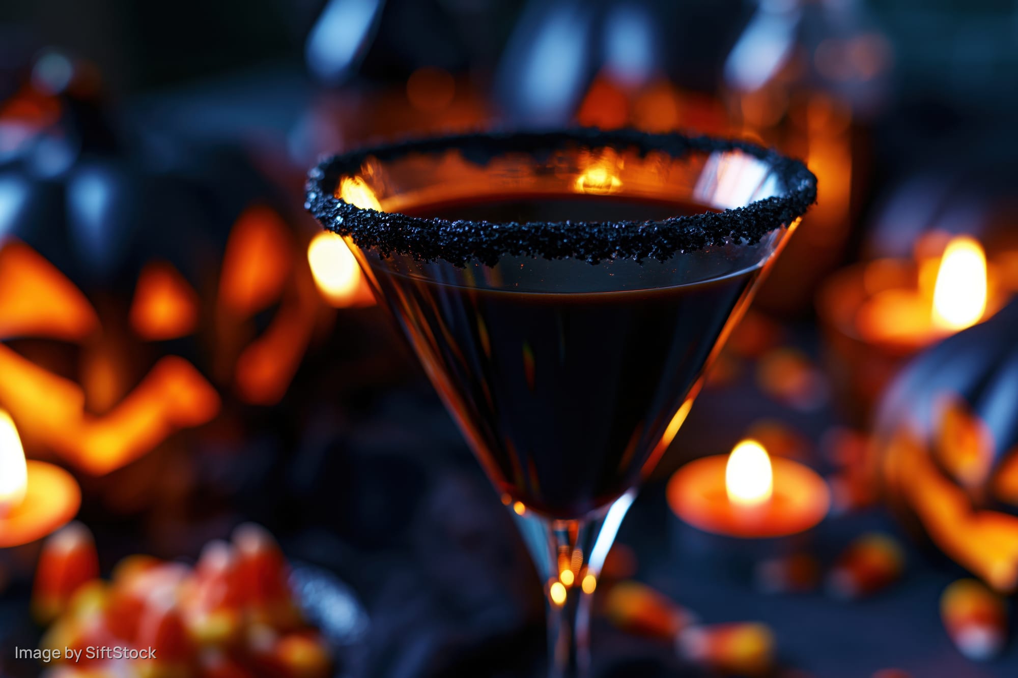 Dark cocktail in a martini glass with sugared black rim surrounded by candles and candy corn on a halloween-themed table