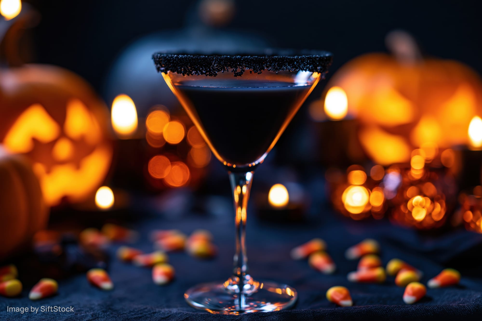 Dark cocktail in a martini glass with sugared black rim on a table surrounded by glowing jack-o-lanterns and candy corn for halloween