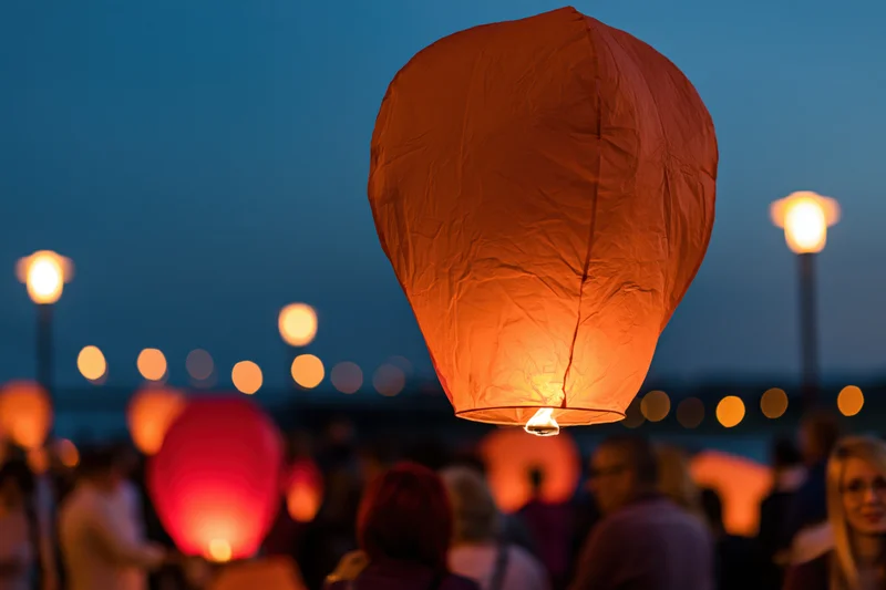 Glowing orange paper sky lantern floating upward at dusk over a crowd with blurred lights in the background