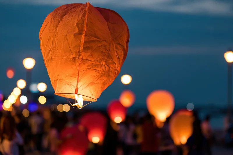 Glowing orange paper sky lantern floating upward at dusk with blurred crowd and lights below