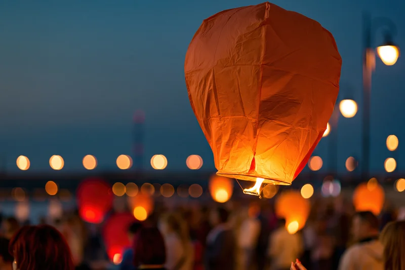 Glowing orange paper sky lantern floating upward at dusk over a crowd